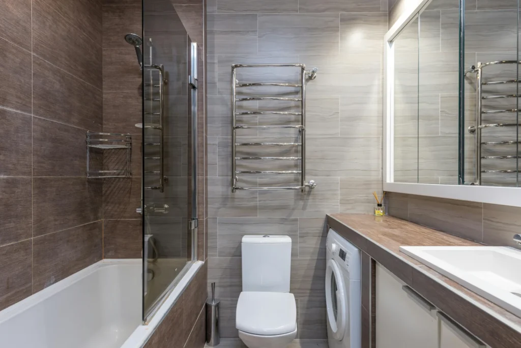 Kleines Badezimmer mit abklappbarer Glas-Duschabtrennung an der Wanne, Heizkörper und Waschmaschine.
